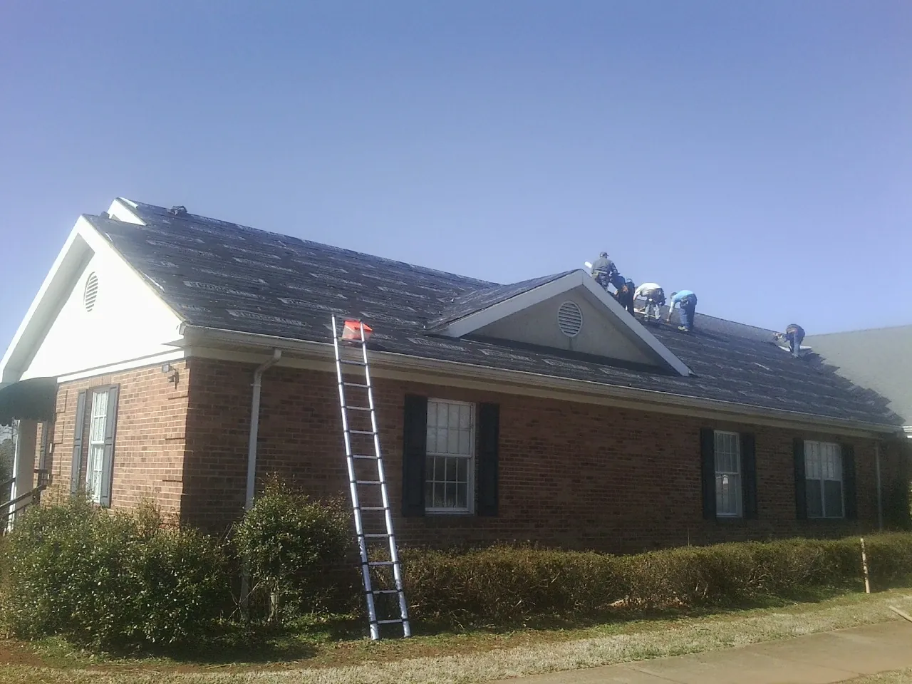 Crew tearing shingles off a brick ranch roof with a ladder set — Emergency Roof Repair work by our crew in Daisetta